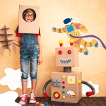 Criança em pé brincando com caixa e um robô de papelão montado ao lado do menino, aludindo à metodologia steam.