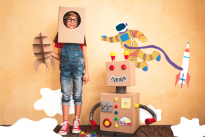 Criança em pé brincando com caixa e um robô de papelão montado ao lado do menino, aludindo à metodologia steam.