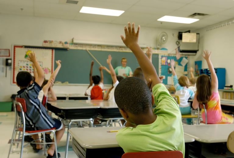 A imagem mostra uma sala de aula com crianças com a mão levantada para responder algo para o professor.