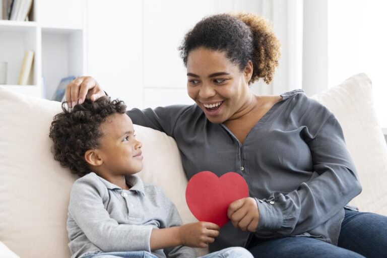 escuta empática: uma mãe negra com seu filho sentados no sofá segurando ambos um coração de papel vermelho.
