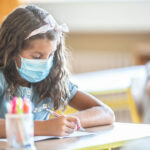 Criança estudando na escola de máscara, refletindo os impactos da pandemia na educação.