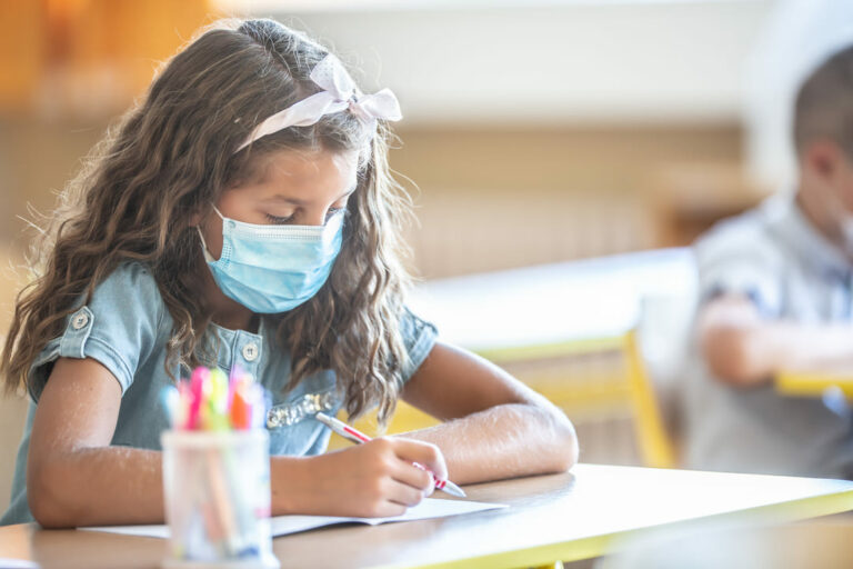 Criança estudando na escola de máscara, refletindo os impactos da pandemia na educação.