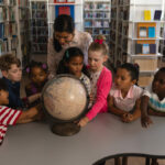 Professora segurando o globo terrestre em biblioteca com alunos em volta, representando o Ensino Religioso no Brasil.