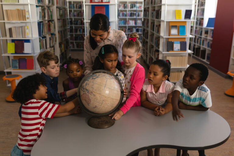 Professora segurando o globo terrestre em biblioteca com alunos em volta, representando o Ensino Religioso no Brasil.